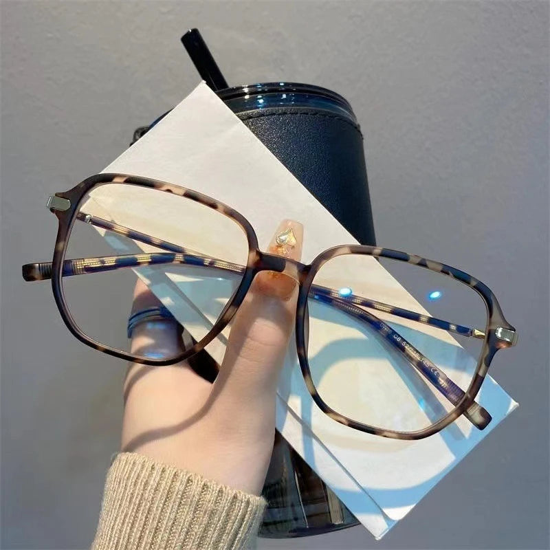 A hand holds a pair of retro anti-blue light sunglasses resting on a white paper next to a dark cup.