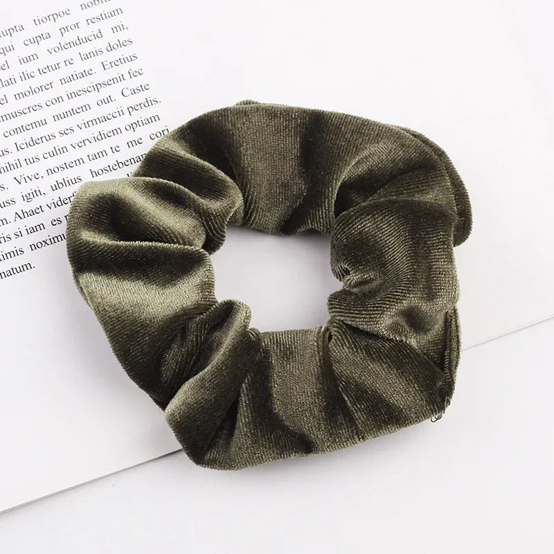 A close-up shot of a dark olive green velvet scrunchie, showing its textured fabric and gathered design.