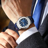 A close-up of a luxury square men's watch with a blue dial and silver casing worn on a wrist, next to a suit jacket.