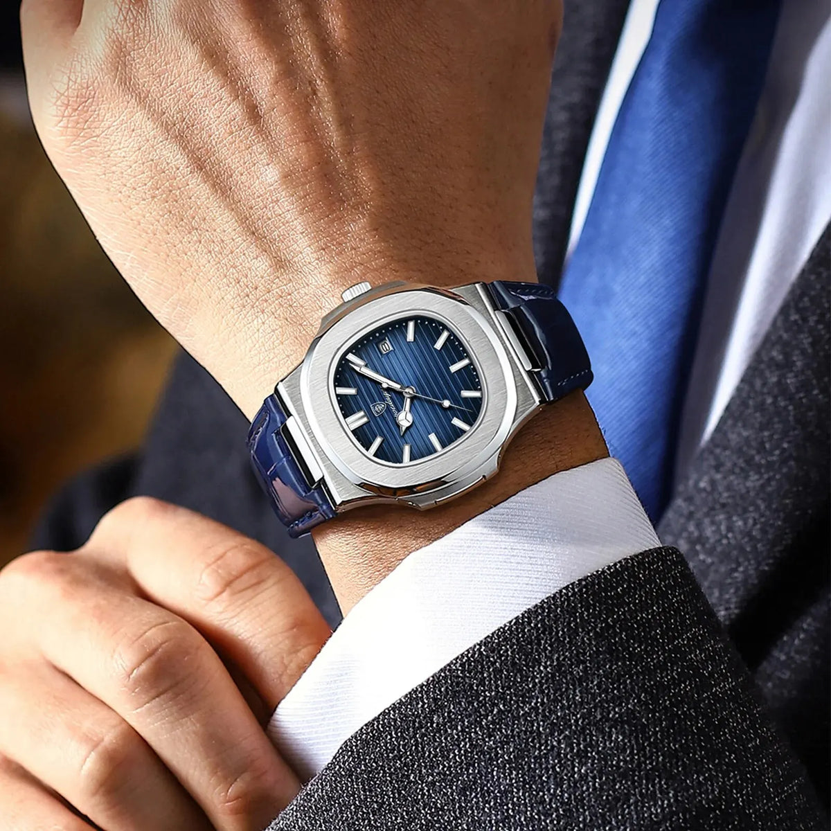 A close-up of a luxury square men's watch with a blue dial and silver casing worn on a wrist, next to a suit jacket.