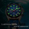 A close-up of a blue and black watch with luminous green hands and a dark face set against a blurred background.