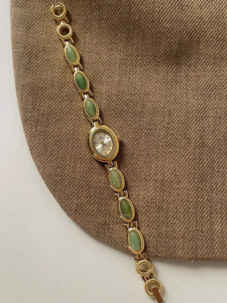 Bracelet en chaîne doré avec des perles de quartz jade vert ovale et un cadran rond, sur fond de tissu marron.