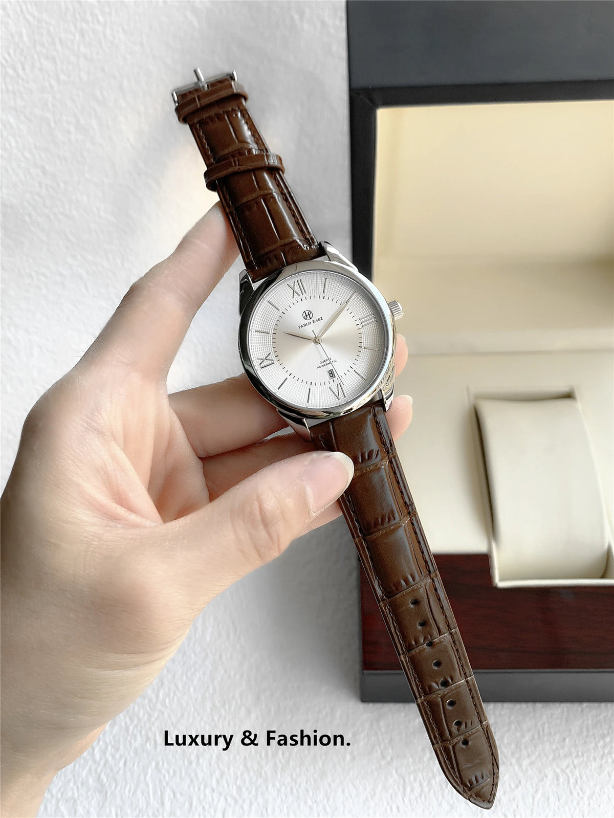 Une main tient une montre-bracelet quartz de luxe pour homme en cuir marron avec un boîtier argenté et un cadran blanc.