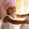 A smiling young girl wearing a pink A-Z letter sleep cap while sitting near a window.