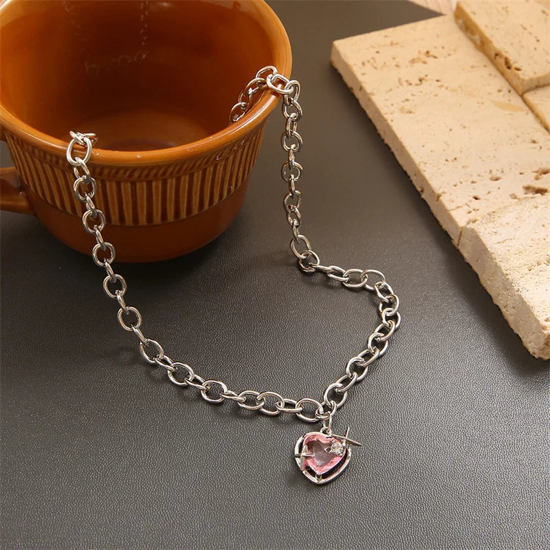 Silver chain necklace with a pink crystal heart pendant resting on a dark surface next to a ceramic cup and bread.