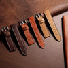 A collection of brown and tan handmade calfskin leather watch straps laid diagonally on a dark brown surface.