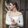 Une femme portant une robe blanche à épaules dénudées et une ceinture dorée, tenant une tige de fleurs séchées.