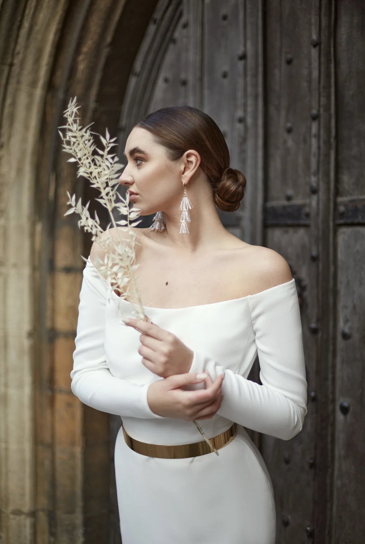 Une femme portant une robe blanche à épaules dénudées et une ceinture dorée, tenant une tige de fleurs séchées.