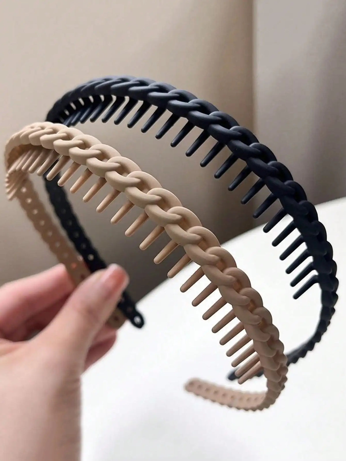 A hand is holding a black braided hair headband and a beige braided hair headband.