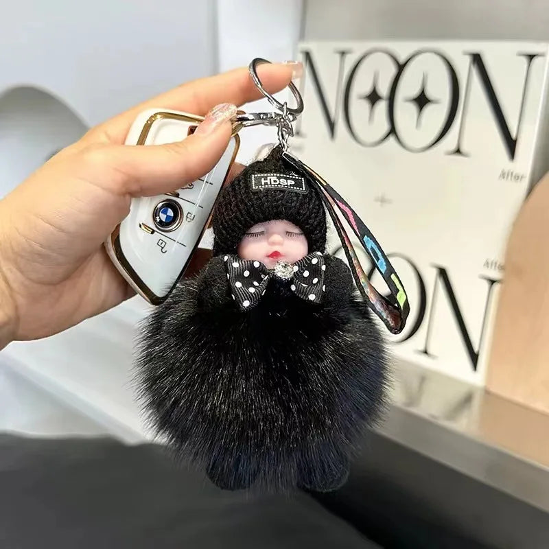 A hand holds a black fluffy plush doll with a bow and a keychain attached to a white and gold object.