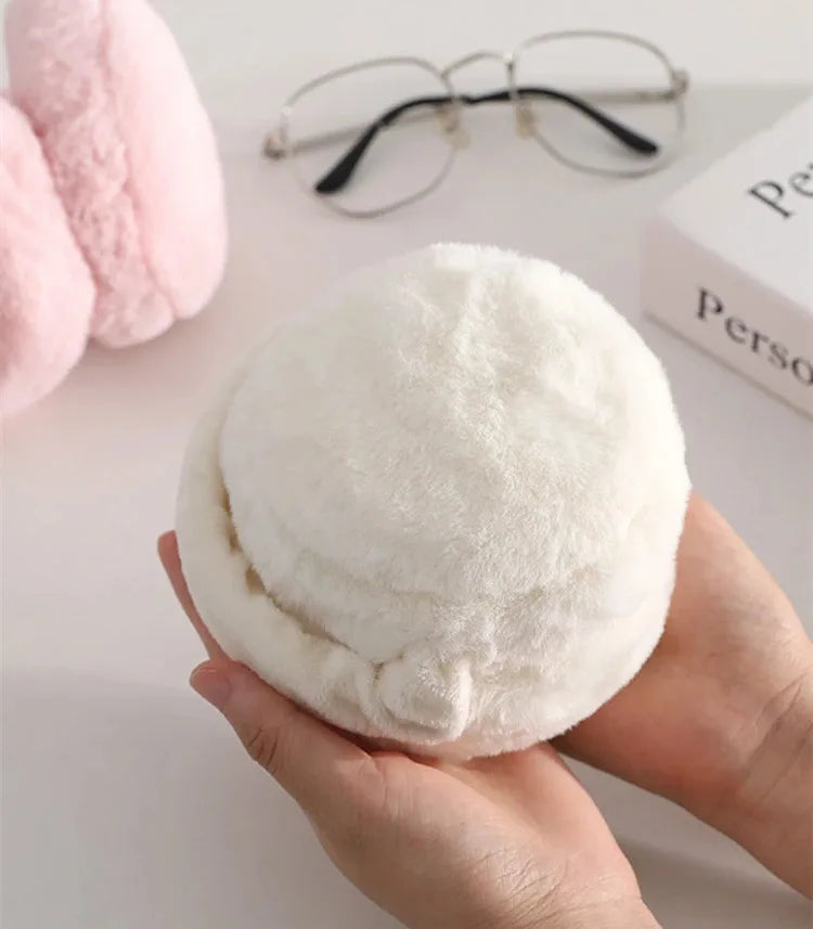 A hand holds a white plush fur ear warmer or earmuff against a light background.