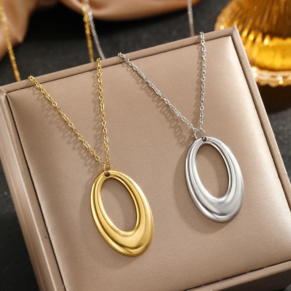 Ein Paar geometischer Halsketten aus Edelstahl, eine goldene und eine silberne, in einer hellbraunen Schachtel präsentiert.
