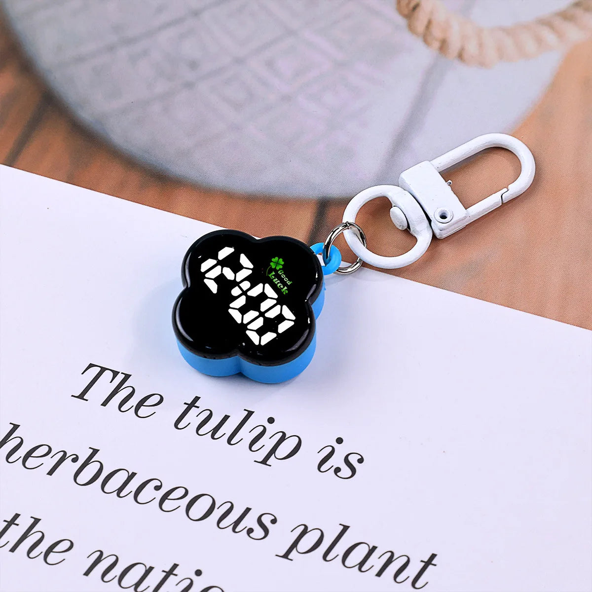 A black LED digital keychain watch with a flower design is clipped to a white paper.
