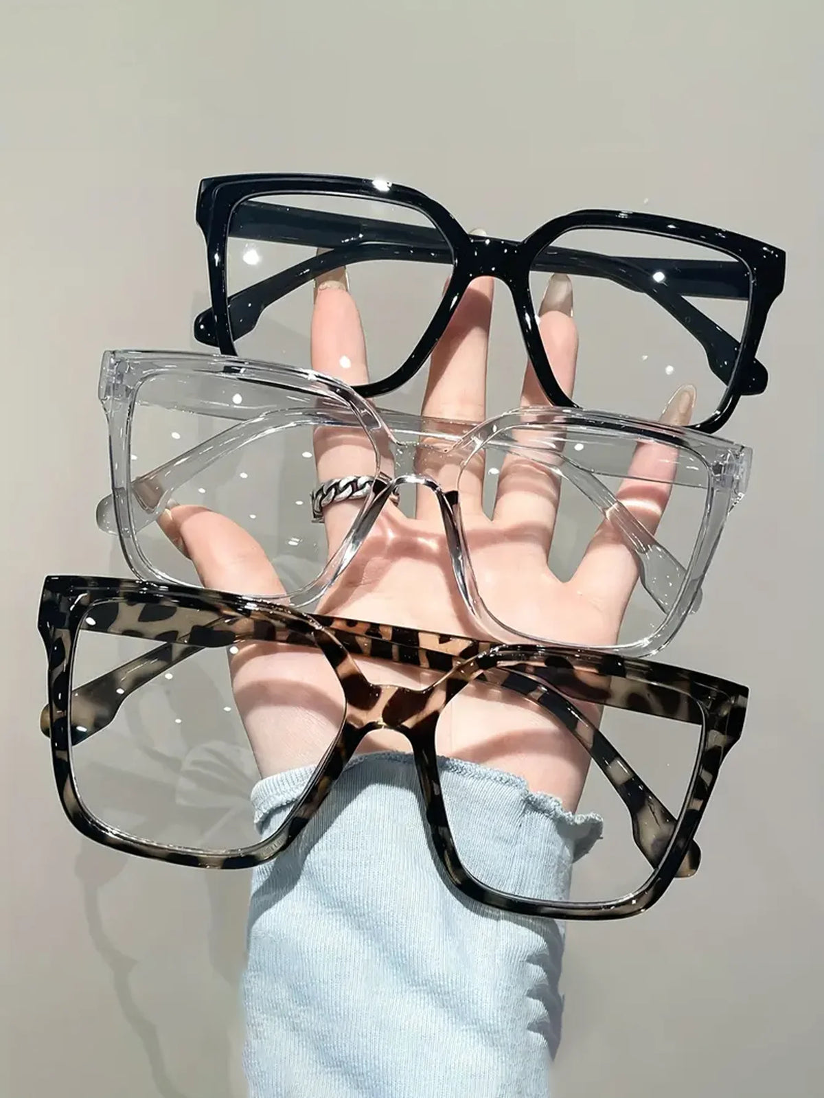 A hand holds a stack of four pairs of elegant geometric reading glasses with various colored frames and transparent lenses.