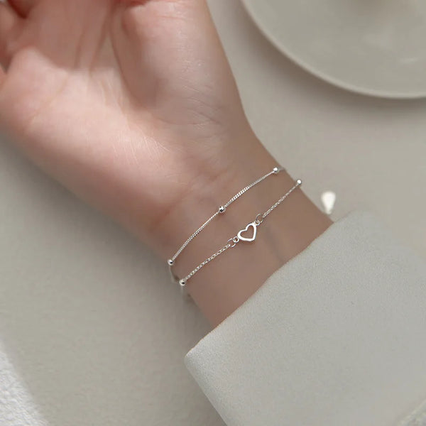 A close-up shot of a hand wearing a sterling silver double heart charm bracelet on a light background.