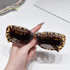 A hand holds a pair of brown leopard print sunglasses with gold accents against a white background.