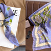 A purple floral print silk scarf draped on a light-colored card next to a brown leather sofa.