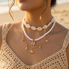 A close-up of a woman wearing a layered gold and white beaded necklace featuring shell and starfish charms.