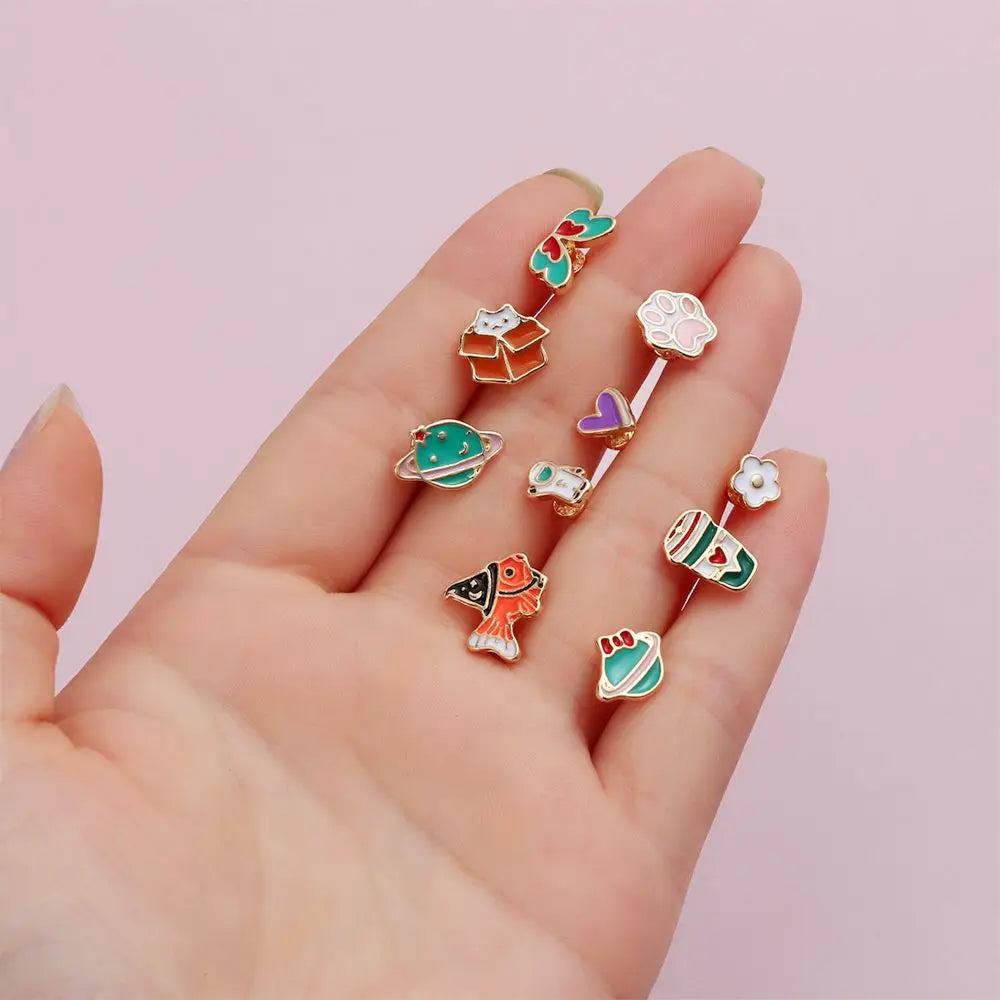 Una mano sostiene varias ornamentos de pulsera de metal decorativos con diseños de esmalte en tonos verde y rosa.