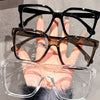 A hand holds a stack of elegant geometric reading glasses with transparent lenses, featuring black, brown, and clear frames.