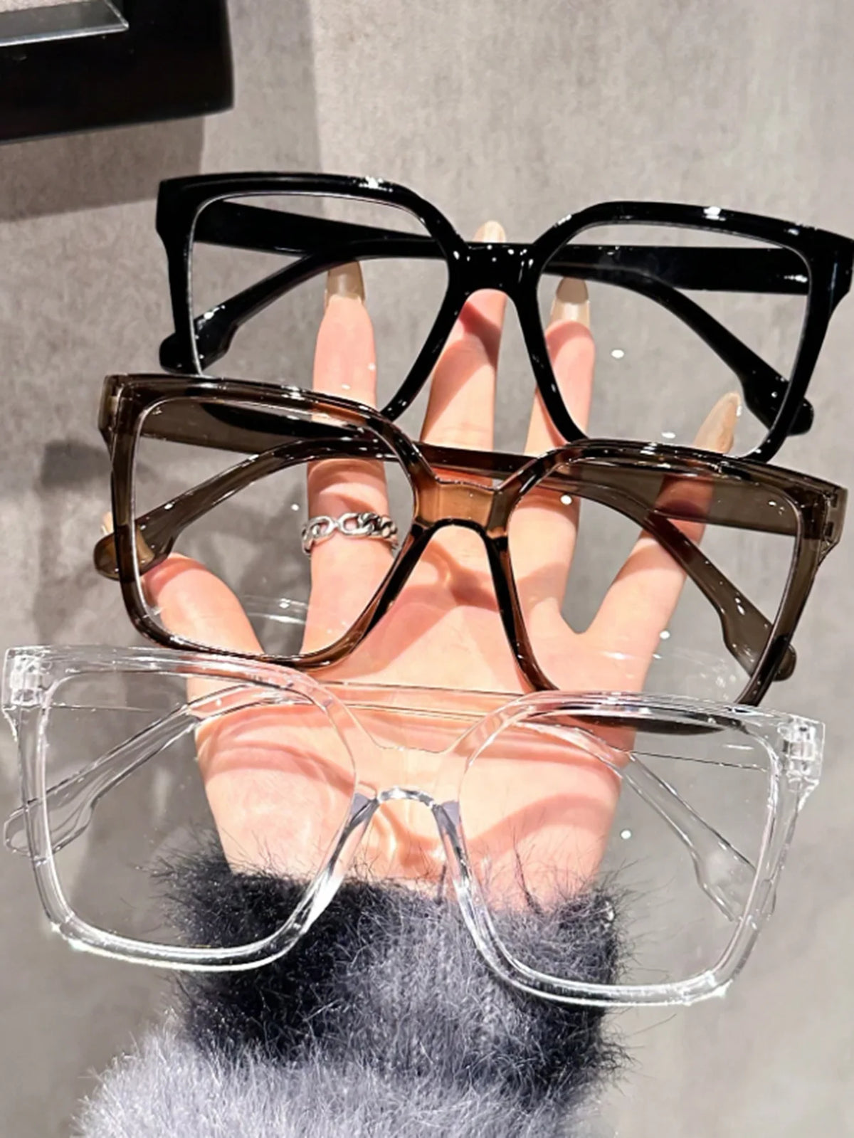 A hand holds a stack of elegant geometric reading glasses with transparent lenses, featuring black, brown, and clear frames.