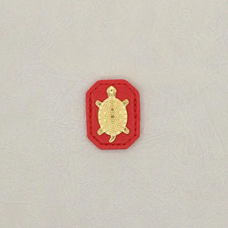 A small, gold-colored turtle charm set within a red, hexagonal-shaped fabric pouch.