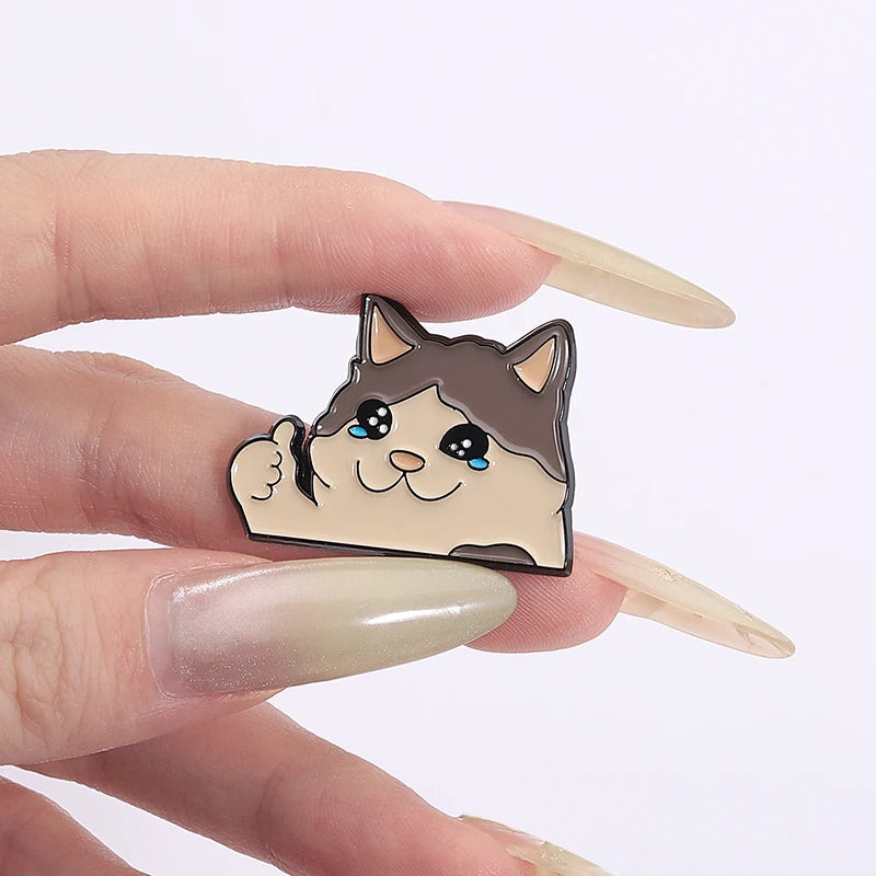 A cute cat crying enamel brooch pin is held between a person's fingers.