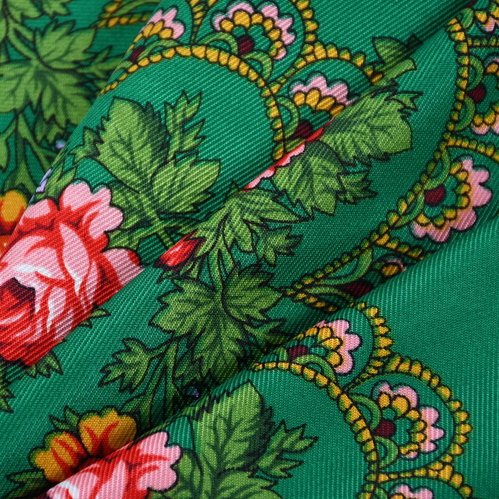 A close-up of a luxury floral print square scarf in shades of green, pink, and yellow on a silk fabric.