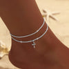 A delicate silver sparkling zircon heart cross anklet worn on a person's ankle against a sandy background.