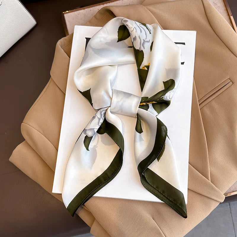 A luxury silk scarf featuring a white and olive green floral pattern tied in a bow, displayed on a white card against a beige