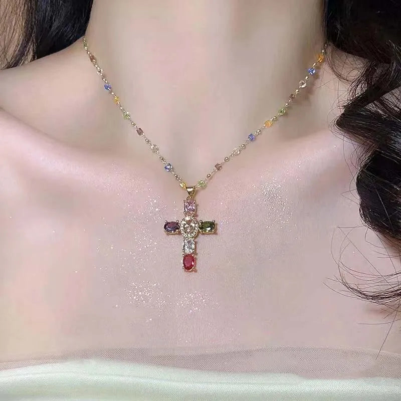A gold chain necklace featuring a colorful zircon cross pendant worn against a light pink fabric background.