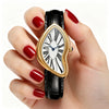 A hand holds a gold-toned quartz watch with an irregular, heart-shaped face and a black strap.