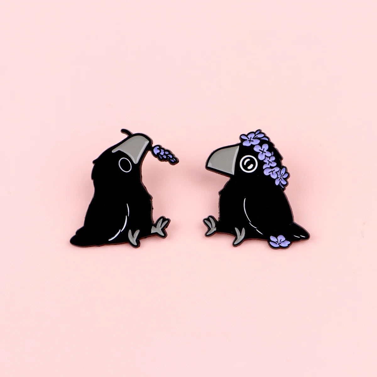 Ein Paar schwarze Email-Anstecker in Form von Krähen mit lila Blumen auf dem Kopf.