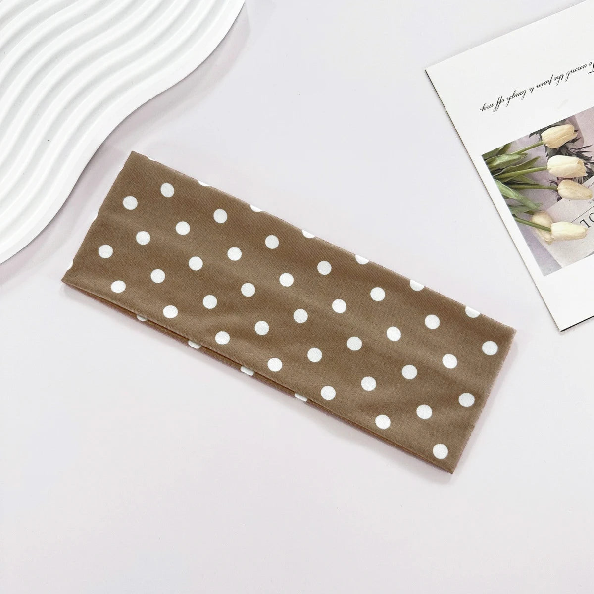 A brown fabric headband with white polka dots is displayed on a white surface next to a magazine.
