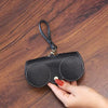 A black leather pouch with two circular compartments and a leather strap is held by a hand on a wooden surface.
