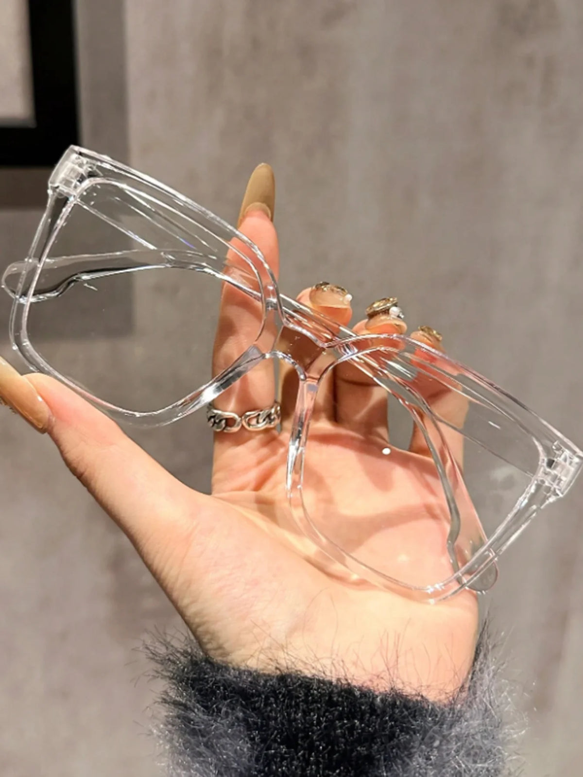 Una mano sostiene un par de gafas cuadradas transparentes contra un fondo gris claro.