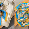A vibrant blue silk scarf with a floral print and gold accents is displayed on a light background.