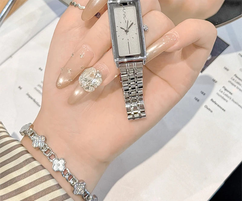 A hand wearing a silver stainless steel quartz watch with a metal bracelet and gold nail polish, resting on a document.
