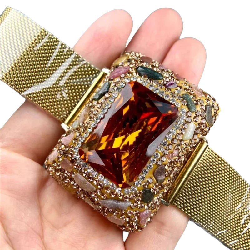 A hand holds a gold-plated swirl cuff armband watch with a square amber stone and textured gold band.