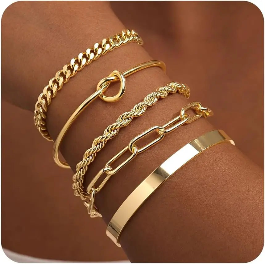 A stack of various gold-toned bangles, including a thick chain bracelet and a thin cuff, worn on a wrist.