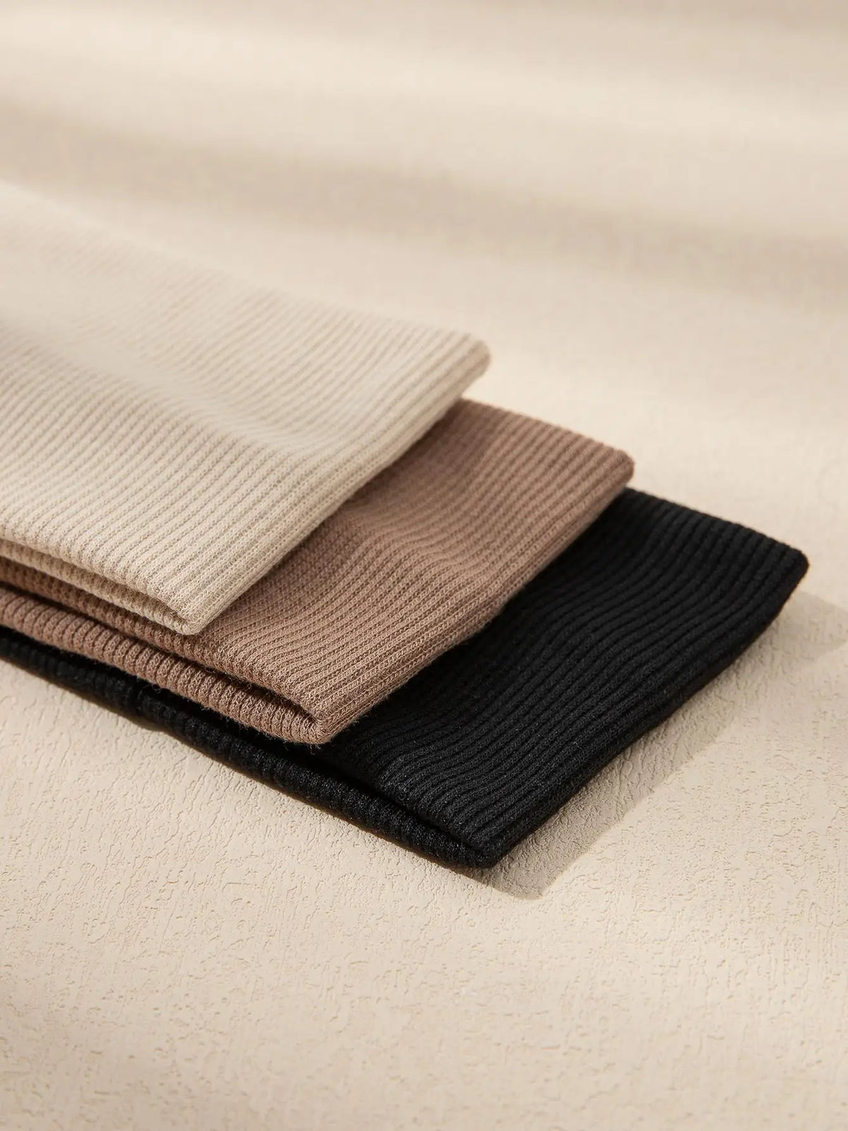 A stack of three solid color fashion headbands in beige, taupe, and black resting on a light background.