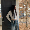 A woman with long dark hair wearing a white shirt and denim jeans stands next to a dark door.