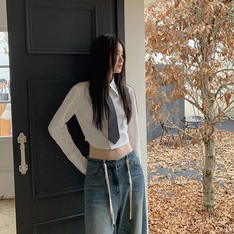 A woman with long dark hair wearing a white shirt and denim jeans stands next to a dark door.