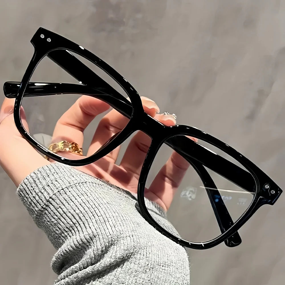 A hand holds a pair of classic black square frames for eye protection against a light gray background.