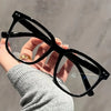A hand holds a pair of classic black square frames for eye protection against a blurred gray background.