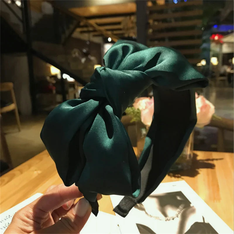 A dark emerald green satin wide bow headband is held up against a blurred indoor background.