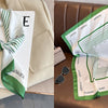A white silk scarf with green stripes is displayed on a white card and a brown leather sofa.