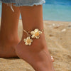 A close-up of a woman's ankle showing a gold floral pendant bracelet and matching earrings against a beach background.