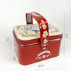 A red tinplate candy box with a carrying loop and floral pattern, shown from the side.
