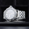 A silver stainless steel watch with a light dial and a metal link band is shown on a dark surface.